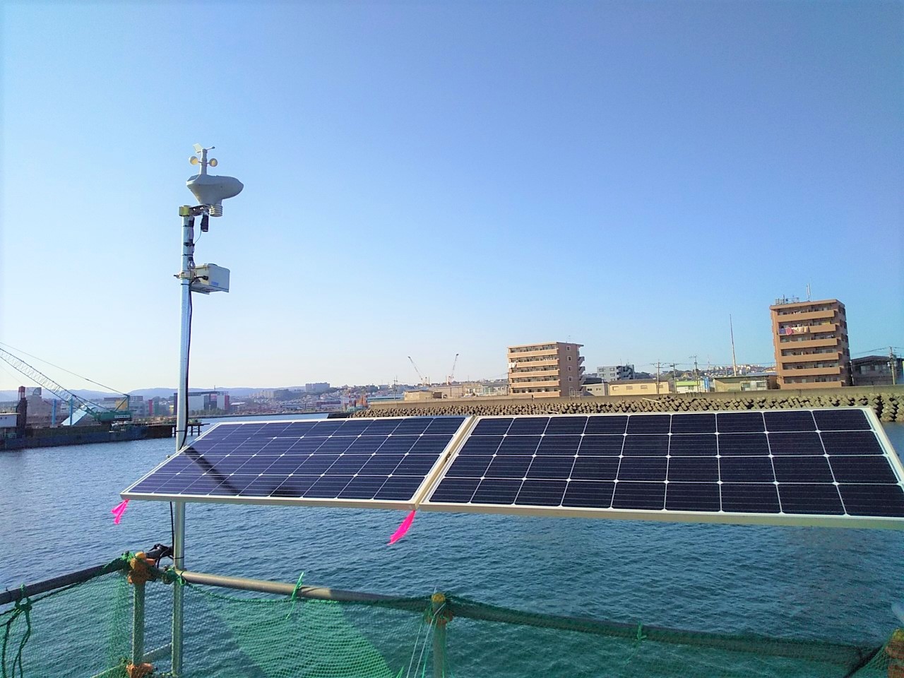 A large, black solar panel on a green fence is mounted on a boat, with a cityscape in the background and a crane in the distance.