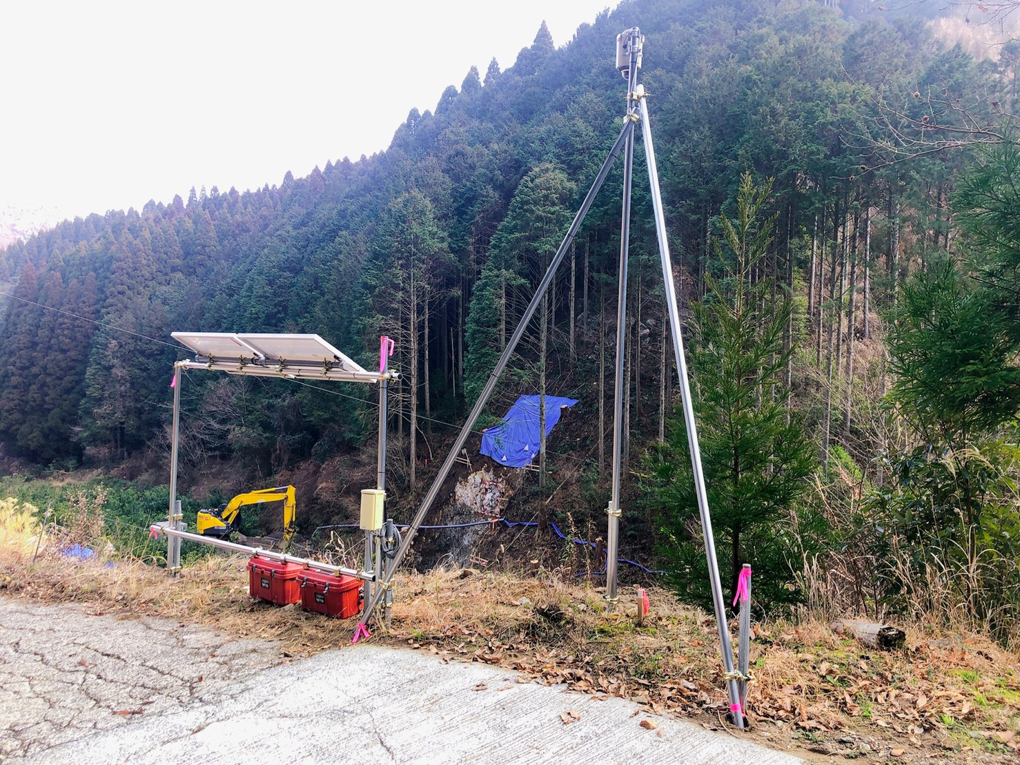 A mountainous landscape with a blue sky, green trees, and a tall metal structure with a camera and yellow crane, surrounded by red and white barriers, is captured in a serene, overcast scene.